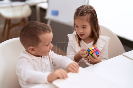 Foto de Adorable girl and boy playing with color puzzle cube sitting on table at kindergarten - Imagen libre de derechos