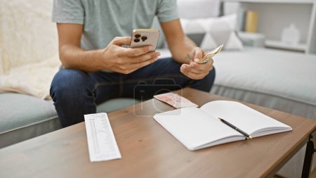 Photo for Young man relaxing on sofa at home, managing his wealth, counting shekels, texting business deals on smartphone - Royalty Free Image
