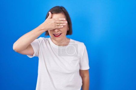 Téléchargez les photos : Middle age hispanic woman standing over blue background smiling and laughing with hand on face covering eyes for surprise. blind concept. - en image libre de droit