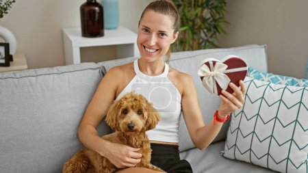 Foto de Joven mujer caucásica con perro sentado en el sofá juntos sosteniendo regalo en casa - Imagen libre de derechos