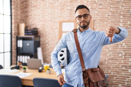 Photo for Young hispanic man working at the office holding bike helmet with angry face, negative sign showing dislike with thumbs down, rejection concept - Royalty Free Image