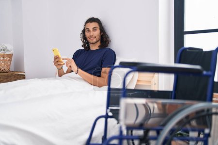 Foto de Young hispanic man disabled using smartphone sitting on bed at bedroom - Imagen libre de derechos