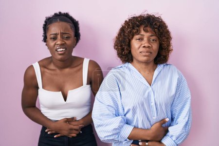Téléchargez les photos : Deux femmes africaines debout sur fond rose avec la main sur le ventre parce que l'indigestion, la maladie douloureuse se sentent mal. concept de douleur. - en image libre de droit