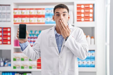 Foto de Hombre hispano guapo que trabaja en la farmacia mostrando la pantalla del teléfono inteligente cubriendo la boca con la mano, sorprendido y asustado por el error. expresión sorprendida - Imagen libre de derechos