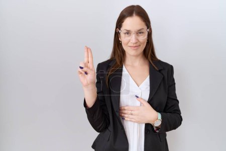 Téléchargez les photos : Belle femme brune portant une veste d'affaires et des lunettes souriantes jurant avec la main sur la poitrine et les doigts vers le haut, faisant un serment de fidélité - en image libre de droit