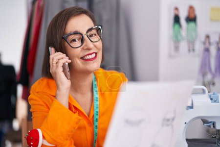 Téléchargez les photos : Middle age woman tailor talking on smartphone looking clothing design at tailor shop - en image libre de droit