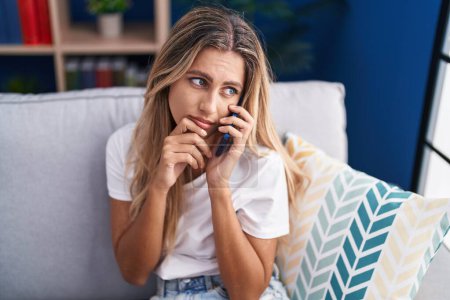 Téléchargez les photos : Jeune femme blonde parlant sur smartphone avec une expression inquiète à la maison - en image libre de droit