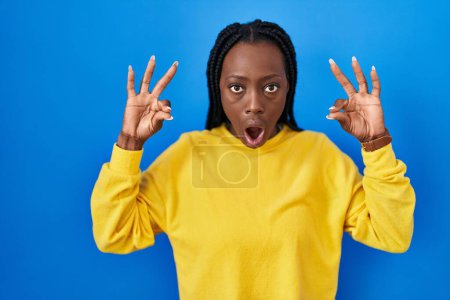 Photo for Beautiful black woman standing over blue background looking surprised and shocked doing ok approval symbol with fingers. crazy expression - Royalty Free Image