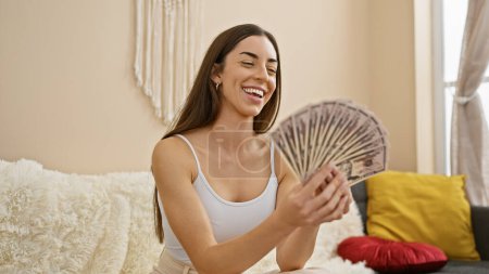 Photo for Attractive young hispanic woman smiling, flaunting her wealth as she holds dollar banknotes, sitting in the comfort of her home's lavish living room sofa. - Royalty Free Image