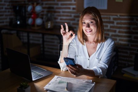 Téléchargez les photos : Jeune rousse femme travaillant au bureau la nuit faire ok signe avec les doigts, sourire geste amical excellent symbole - en image libre de droit