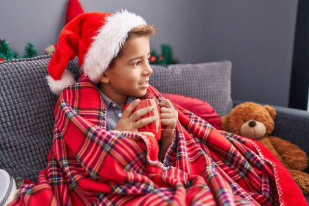 Foto de Adorable niño hispano bebiendo leche sentado en el sofá por la decoración de Navidad en casa - Imagen libre de derechos