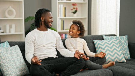 Téléchargez les photos : Afro-américain père et fille assis sur canapé faire de l'exercice de yoga à la maison - en image libre de droit