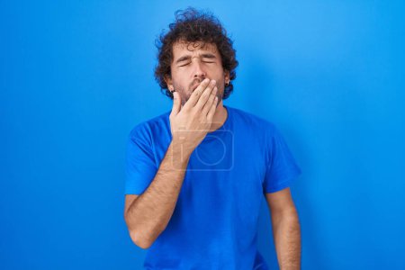 Foto de Joven hispano parado sobre fondo azul aburrido bostezando cansado cubriendo la boca con la mano. inquietud y somnolencia. - Imagen libre de derechos