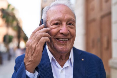 Téléchargez les photos : Moyen âge homme aux cheveux gris souriant confiant parler sur le smartphone à la rue - en image libre de droit