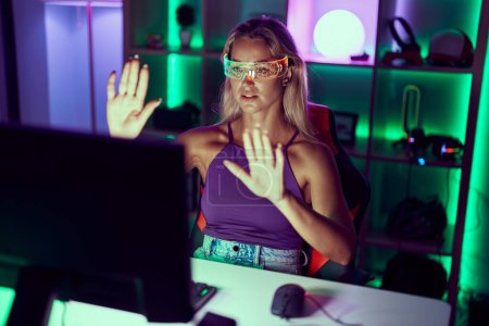 Foto de Joven mujer rubia streamer jugando videojuego utilizando gafas de realidad virtual en la sala de juegos - Imagen libre de derechos