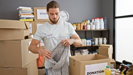 Photo for Young hispanic man volunteer packing clothes on cardboard box at charity center - Royalty Free Image