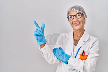 Photo for Middle age woman with grey hair wearing scientist robe with a big smile on face, pointing with hand and finger to the side looking at the camera. - Royalty Free Image