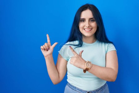 Photo for Young modern girl with blue hair standing over blue background smiling and looking at the camera pointing with two hands and fingers to the side. - Royalty Free Image