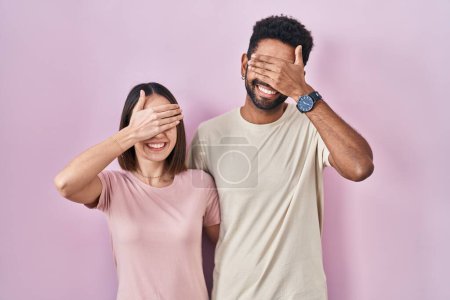 Foto de Jóvenes pareja hispana juntos sobre fondo rosa sonriendo y riendo con la mano en la cara cubriendo los ojos para sorpresa. concepto ciego. - Imagen libre de derechos