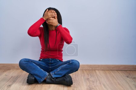 Téléchargez les photos : Jeune afro-américain avec des tresses assis sur le sol à la maison couvrant les yeux et la bouche avec les mains, surpris et choqué. cacher l'émotion - en image libre de droit