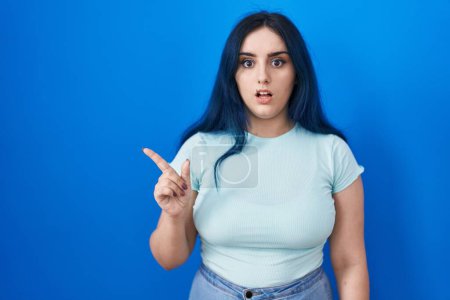 Photo for Young modern girl with blue hair standing over blue background surprised pointing with finger to the side, open mouth amazed expression. - Royalty Free Image
