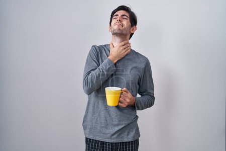 Foto de Hombre hispano joven usando pijama bebiendo una taza de café tocando el cuello doloroso, dolor de garganta por gripe, clod e infección - Imagen libre de derechos
