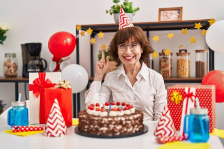 Foto de Mujer de mediana edad celebrando cumpleaños sosteniendo gran pastel de chocolate sorprendido con una idea o pregunta señalando el dedo con la cara feliz, número uno - Imagen libre de derechos