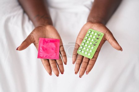 Foto de Mujer afroamericana sosteniendo condón y píldoras anticonceptivas en el dormitorio - Imagen libre de derechos