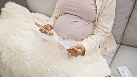 Téléchargez les photos : Nichée dans une couverture à la maison, une jeune femme enceinte, chaudement assise sur son canapé, se livre à des devoirs de maternité tout en lisant des documents importants pour bébé à l'aide de sa technologie de smartphone fidèle. - en image libre de droit