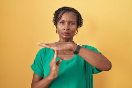 Photo for African woman with dreadlocks standing over yellow background doing time out gesture with hands, frustrated and serious face - Royalty Free Image