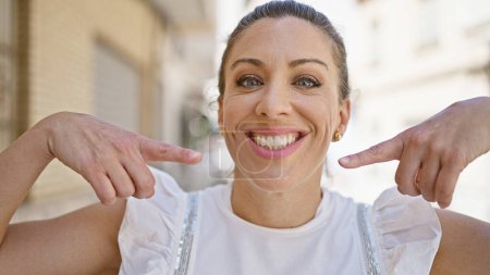 Téléchargez les photos : Jeune femme blonde souriante confiante pointant vers la bouche dans la rue - en image libre de droit