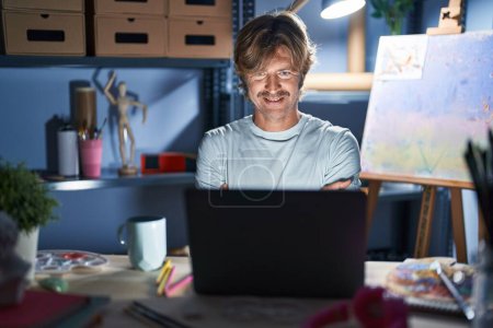 Téléchargez les photos : Homme d'âge moyen assis au studio d'art avec ordinateur portable la nuit visage heureux souriant avec les bras croisés regardant la caméra. personne positive. - en image libre de droit