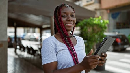 Téléchargez les photos : Femme afro-américaine confiante, rayonnante de bonheur, profitant de son pavé tactile sur une terrasse animée du café, se prélassant dans l'ambiance urbaine énergique de la ville. - en image libre de droit
