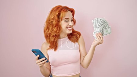 Photo for Young redhead woman smiling holding dollars and smartphone over isolated pink background - Royalty Free Image