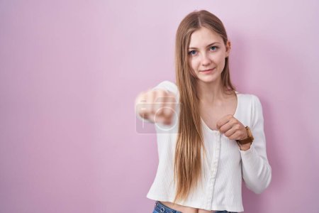 Photo for Young caucasian woman standing over pink background punching fist to fight, aggressive and angry attack, threat and violence - Royalty Free Image