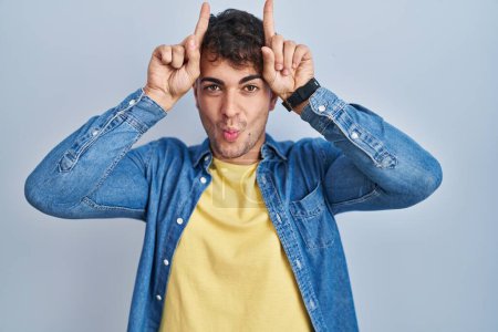 Téléchargez les photos : Jeune homme hispanique debout sur fond bleu faisant un geste drôle avec le doigt sur la tête comme des cornes de taureau - en image libre de droit