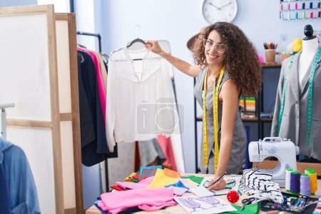 Téléchargez les photos : Jeune belle femme hispanique tailleur souriant confiant tenant t-shirt écriture sur ordinateur portable à l'usine de vêtements - en image libre de droit