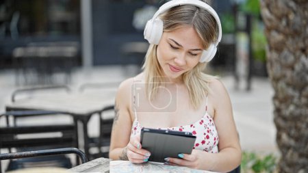 Téléchargez les photos : Jeune femme blonde utilisant touchpad et écouteurs assis sur la table à la terrasse du café - en image libre de droit