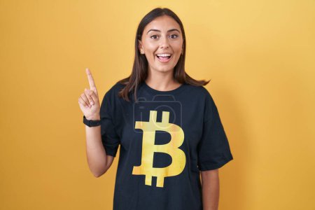 Foto de Mujer hispana joven con camiseta bitcoin sonriendo asombrada y sorprendida y señalando con los dedos y los brazos levantados. - Imagen libre de derechos