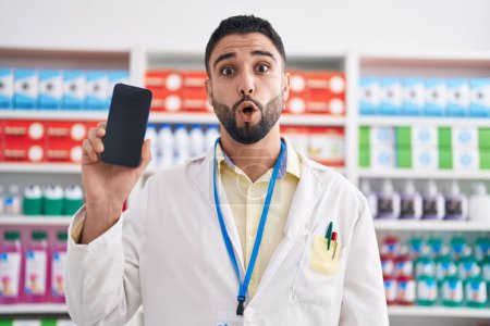 Photo for Hispanic young man working at pharmacy drugstore showing smartphone screen scared and amazed with open mouth for surprise, disbelief face - Royalty Free Image