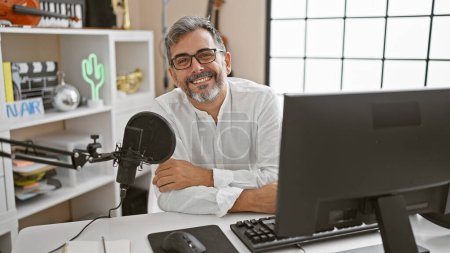 Foto de Joven, atractivo y canoso hombre hispano sonriendo y presentando magistralmente noticias en un programa de radio en vivo dentro de un estudio de radio profesional - Imagen libre de derechos