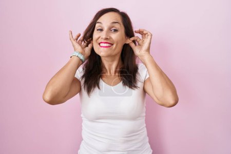 Photo for Middle age brunette woman standing over pink background smiling pulling ears with fingers, funny gesture. audition problem - Royalty Free Image