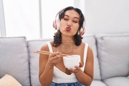 Foto de Joven mujer hispana comiendo comida asiática usando palillos mirando a la cámara soplando un beso siendo encantadora y sexy. expresión de amor. - Imagen libre de derechos