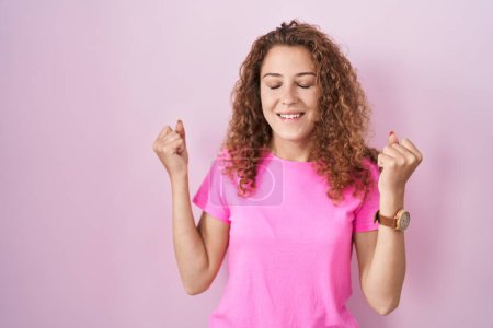 Téléchargez les photos : Jeune femme caucasienne debout sur fond rose excité pour le succès avec les bras levés et les yeux fermés célébrant la victoire en souriant. concept gagnant. - en image libre de droit
