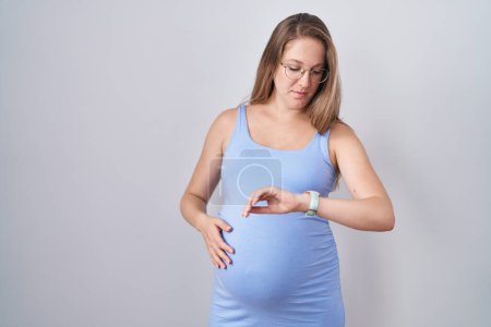 Foto de Mujer embarazada joven de pie sobre fondo blanco comprobar la hora en reloj de pulsera, relajado y seguro de sí mismo - Imagen libre de derechos