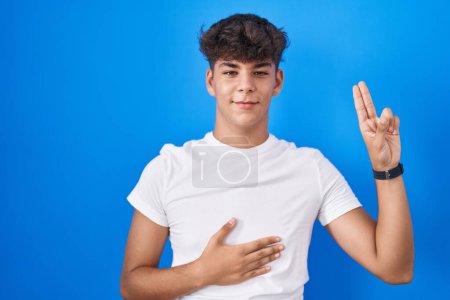 Téléchargez les photos : Adolescent hispanique debout sur fond bleu souriant jurer avec la main sur la poitrine et les doigts vers le haut, faisant un serment de fidélité promesse - en image libre de droit