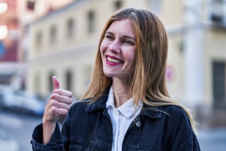 Téléchargez les photos : Jeune femme blonde souriante confiante faire ok signe avec le pouce vers le haut à la rue - en image libre de droit