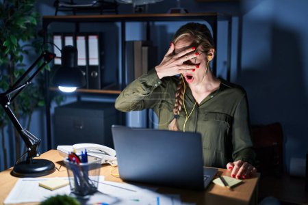 Téléchargez les photos : Young blonde woman working at the office at night peeking in shock covering face and eyes with hand, looking through fingers with embarrassed expression. - en image libre de droit