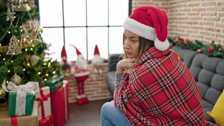 Téléchargez les photos : Jeune femme caucasienne couverte d'une couverture célébrant Noël avec une expression triste à la maison - en image libre de droit