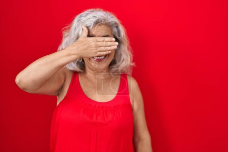 Photo for Middle age woman with grey hair standing over red background smiling and laughing with hand on face covering eyes for surprise. blind concept. - Royalty Free Image